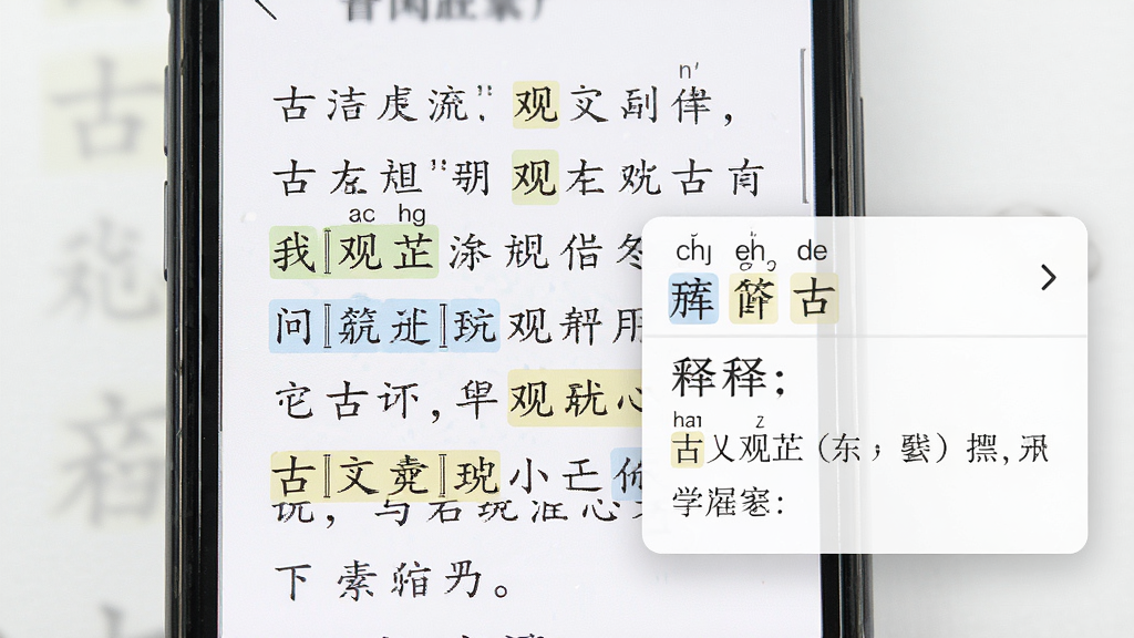 一幅展示古文观芷注释功能的特写图片,画面中央是手机屏幕,屏幕上显示一段古文,其中重点字词被高亮标注,旁边弹出注释框,布局为上下结构,风格为学术清新,细节有拼音标签和释义文字,画质高清,干净,无多余元素