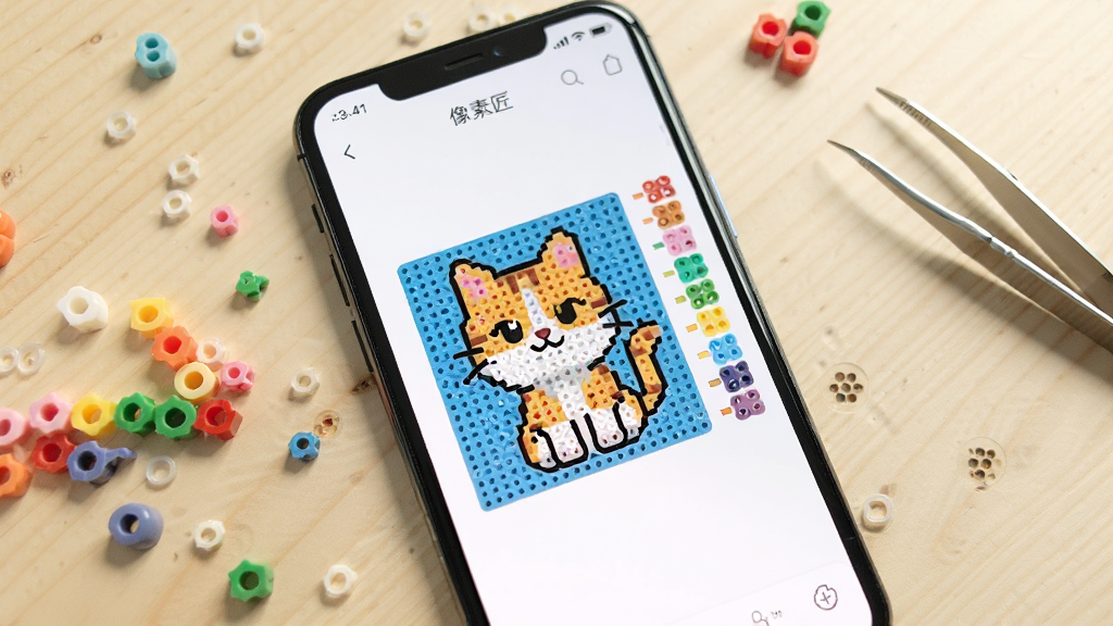 主图展示一部智能手机屏幕,屏幕中打开「像素匠」应用界面,界面中央是一幅正在转换的卡通猫咪拼豆图纸,图纸旁边排列着彩色拼豆颗粒和色号标签。背景是干净的原木桌面,桌面上散落几颗彩色拼豆和一把镊子。整体风格清新明亮,画面简洁,画质清晰,无多余元素。