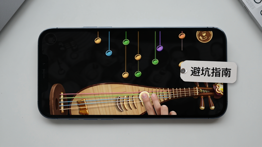 画面展示一部手机横屏运行琵琶曲游戏网页版,屏幕上方有音符掉落,下方为琵琶琴弦,手指正在触击屏幕,旁边有“避坑指南”四个字的标签浮窗,背景为简洁的浅灰色桌面,风格写实清晰,画质干净,无多余元素