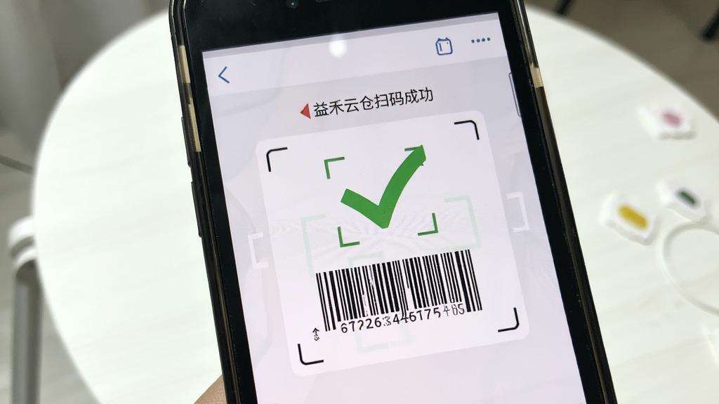 手机镜头对准商品条形码,屏幕显示益禾云仓扫码成功界面,画面中有绿色对勾标识,背景为简洁的白色桌面,风格清新,细节清晰,画质干净,无多余元素
