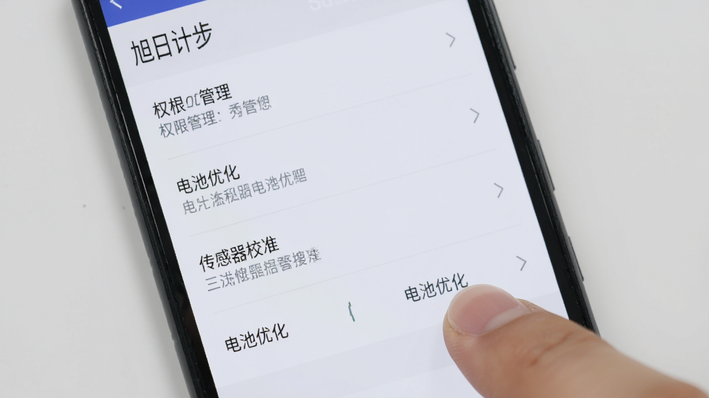 一部安卓手机显示“旭日计步”的设置界面,屏幕上列出了权限管理、电池优化和传感器校准等选项,用户的手指正点击“电池优化”按钮,画面背景为浅灰色,风格简洁直观,画质清晰干净,突出操作步骤