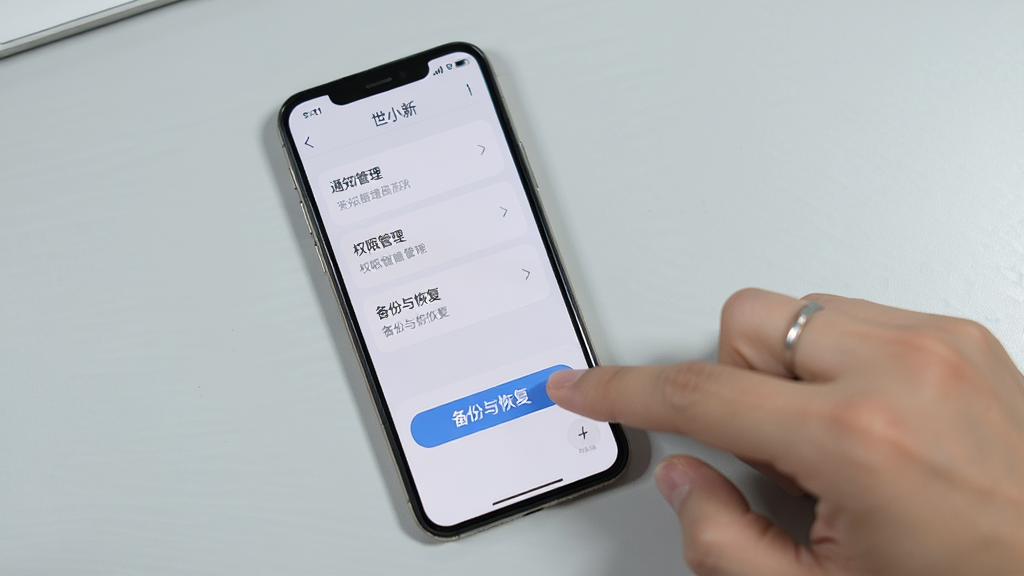 手机屏幕显示“世小新”设置页面,页面中包含通知管理、权限管理、备份与恢复等选项,旁边有一只手正在点击“备份与恢复”按钮。背景为浅灰色桌面,风格写实清晰,画质干净,无多余元素。