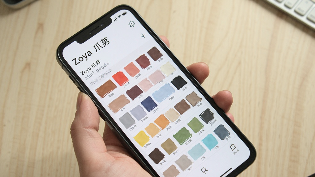 一张展示Zoya爪芽应用在手机上正常运行并显示色卡列表的界面截图,画面主体为手持手机,屏幕内为Zoya爪芽主界面,背景为简洁的浅色桌面,布局采用斜角拍摄,风格生活化,细节包括手指握持手机的自然姿态与屏幕内色块的清晰显示,画质清晰,干净,无多余元素
