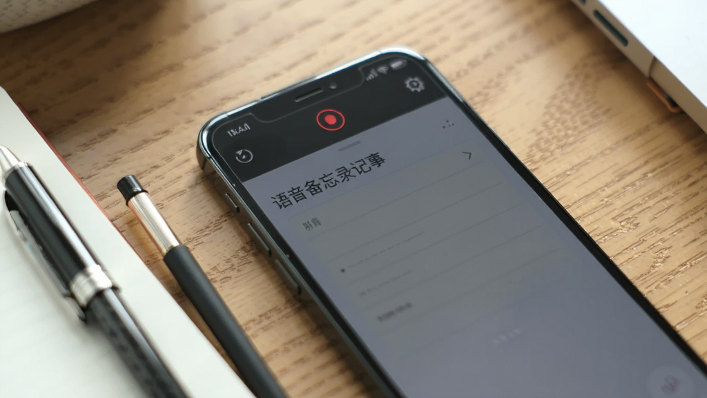 一副手机放在桌面上的场景图,手机屏幕处于息屏状态但顶部状态栏显示录音中的红色指示图标,旁边有一支笔和笔记本,背景为木质桌面,画面风格写实生活化,手机屏幕上隐约可见“语音备忘录记事”字样,光线柔和,画质清晰,无多余杂物