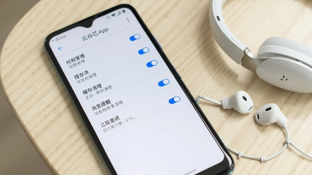 一部安卓手机屏幕显示云谷芯App的设置页面,页面中包含权限管理、缓存清理、消息提醒等选项列表,界面风格简洁,以白色背景和蓝色开关按钮为主,手机平放在浅色桌面上,旁边放着一副白色耳机,整体光线明亮柔和,画质清晰干净,无多余元素