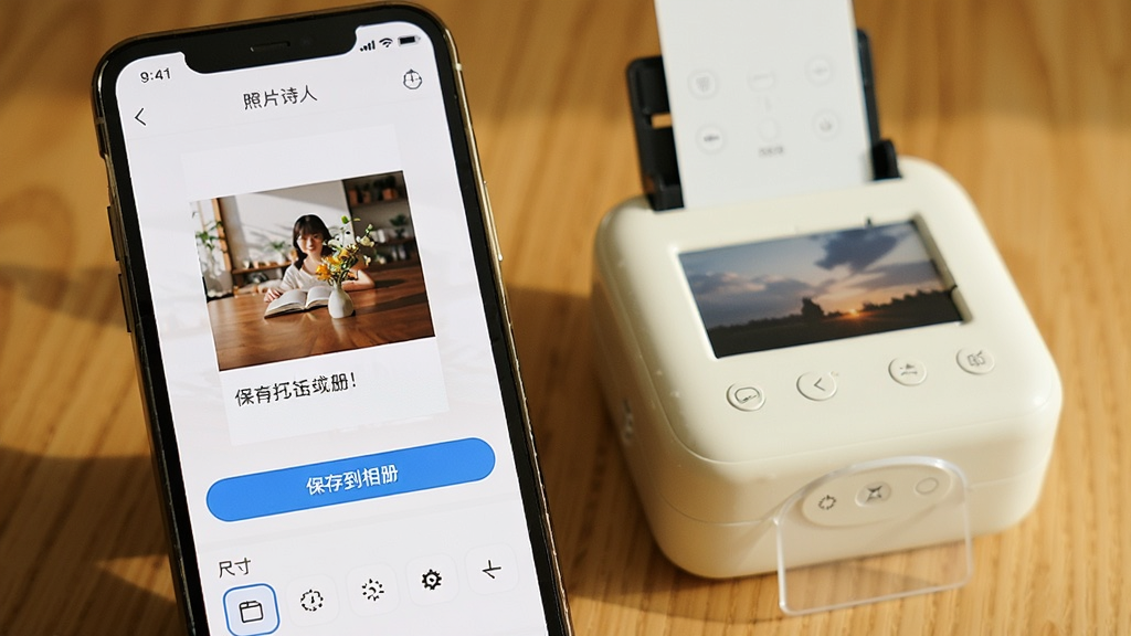 手机屏幕显示“照片诗人”App的导出设置界面,屏幕中有一张配文完成的照片预览图,下方有“保存到相册”按钮和尺寸选择选项,手机旁边放着一台小型照片打印机,整体画面温馨实用,光线明亮,画质清晰,无杂乱背景