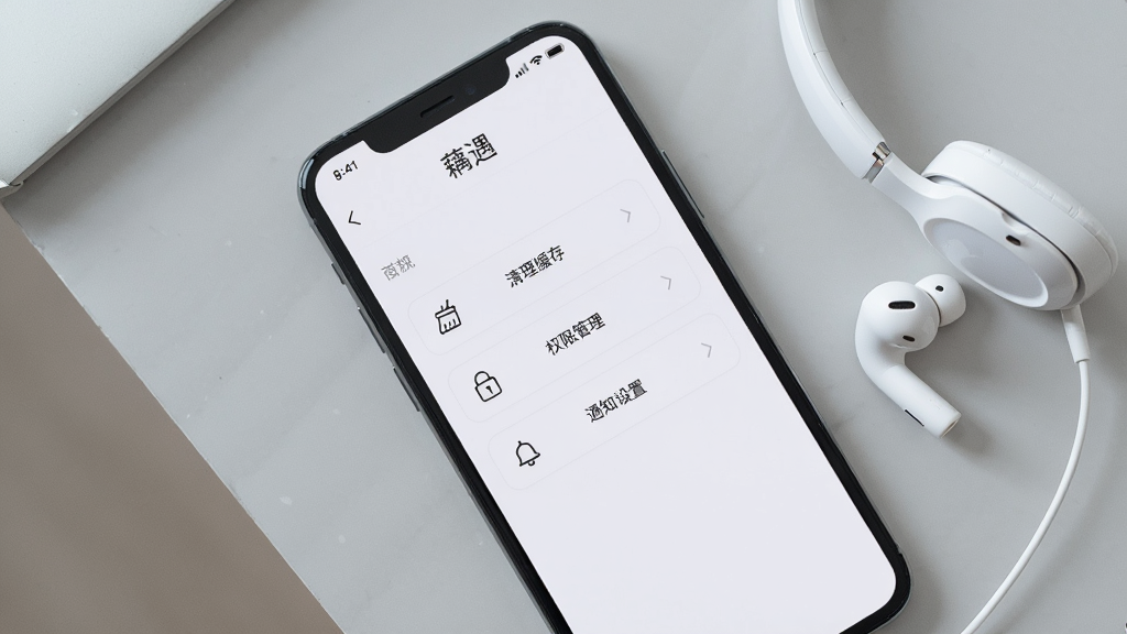 画面主体为一部手机屏幕显示藕遇应用的设置页面,页面中包含“清理缓存”“权限管理”“通知设置”等选项按钮;手机平放在浅灰色桌面上,旁边放着一副白色耳机;布局为俯视平铺构图;风格为简洁数码风;细节上屏幕显示清晰,按钮图标为线性风格;画质为高清产品图,干净无多余元素