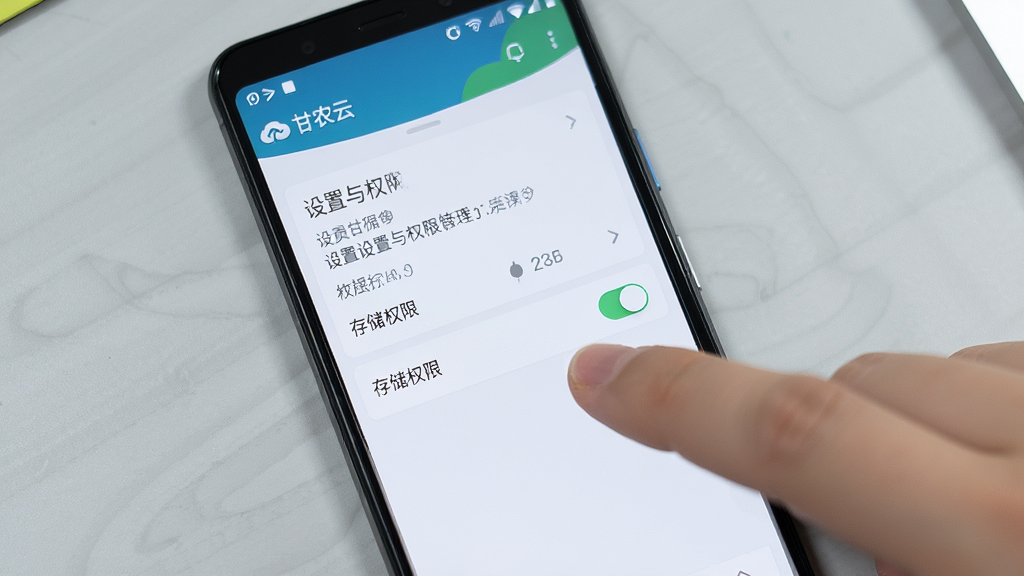 画面主体为一部安卓手机屏幕显示“甘农云”应用设置与权限管理界面,旁边有一只手正点击“存储权限”开关;布局为手机特写居中,背景为浅灰色桌面;风格为简洁数码教程风;细节上屏幕内容清晰,手指动作自然,光线均匀;画质高清,画面干净无杂乱元素