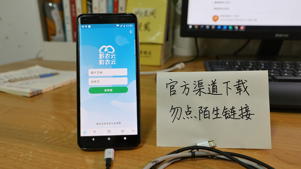 一部安卓手机屏幕显示“黔农云”登录页面,旁边放着一张手写便签写着“官方渠道下载”与“勿点陌生链接”,手机下方有充电线,背景为居家桌面,风格生活化写实,光线明亮,画质清晰干净,突出安全提示氛围