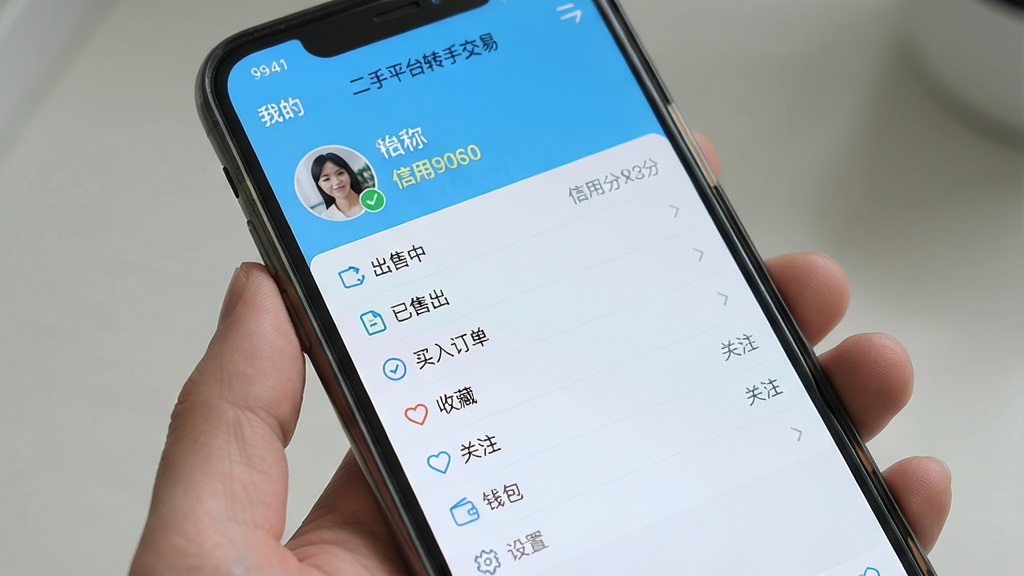 主体为一部手机屏幕,屏幕中显示“二手平台转手交易”的“我的”页面,页面顶部有用户头像、昵称、信用分,下方功能列表包括“出售中”“已售出”“买入订单”“收藏”“关注”“钱包”“设置”等,背景为浅蓝色,布局简洁,风格清爽,细节丰富,画质清晰,干净,无多余元素