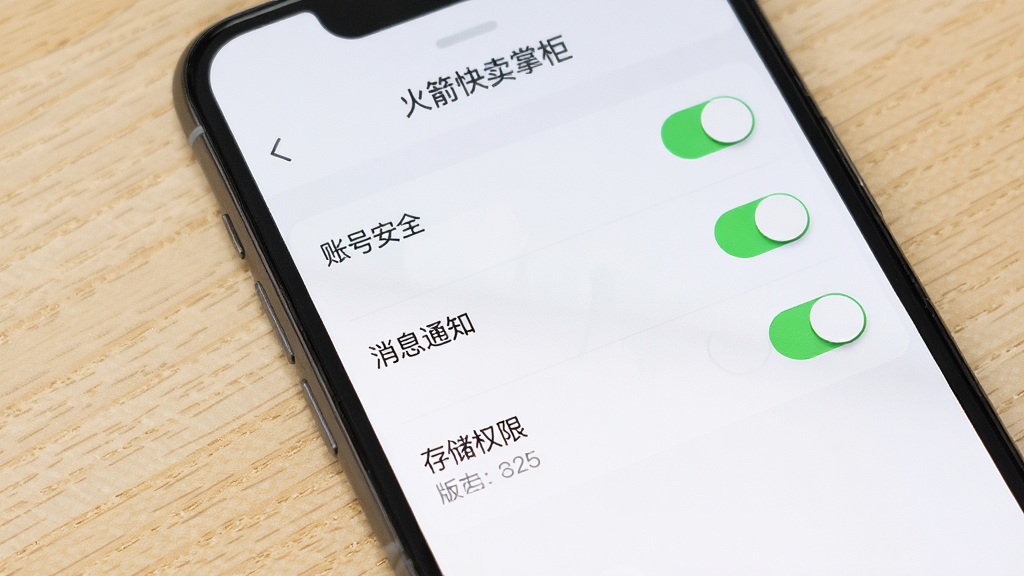 画面主体为智能手机屏幕显示火箭快卖掌柜的设置页面,页面中有“账号安全”、“消息通知”、“存储权限”等选项开关,背景为浅色木纹桌面,布局为列表式,排版简洁,风格实用,细节包括开关按钮和版本号信息,画质高清,干净无多余元素