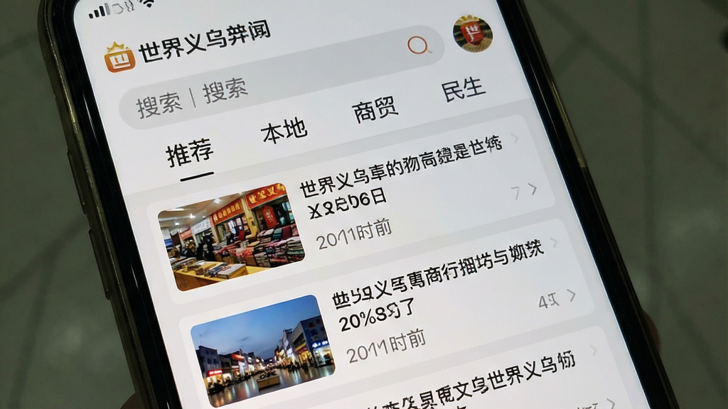 一张手机屏幕截图样式的图片,屏幕中展示“世界义乌”APP的资讯频道,上方为搜索栏和分类标签如“推荐”、“本地”、“商贸”、“民生”,下方为新闻卡片列表,每张卡片包含缩略图、标题和发布时间。布局采用列表式排版,清晰有序。风格为写实屏幕截图风格,背景为浅灰色,模拟手机界面效果。细节上突出“世界义乌”品牌标识和资讯内容,文字清晰可读,画质干净,无多余元素。