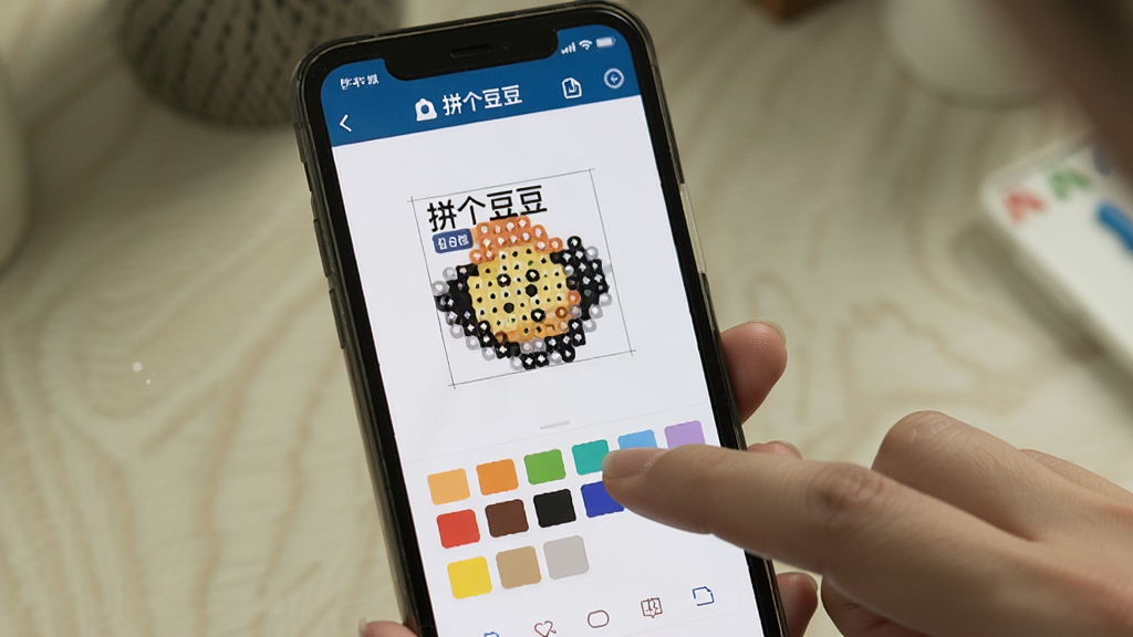 一张展示拼个豆豆软件在手机上运行的实拍照片,主体为手机屏幕显示拼豆图纸编辑界面,布局为竖屏,排版清晰,风格为真实场景拍摄,细节显示手指点击色板操作,画质高清,干净无多余元素,文字标注“拼个豆豆”