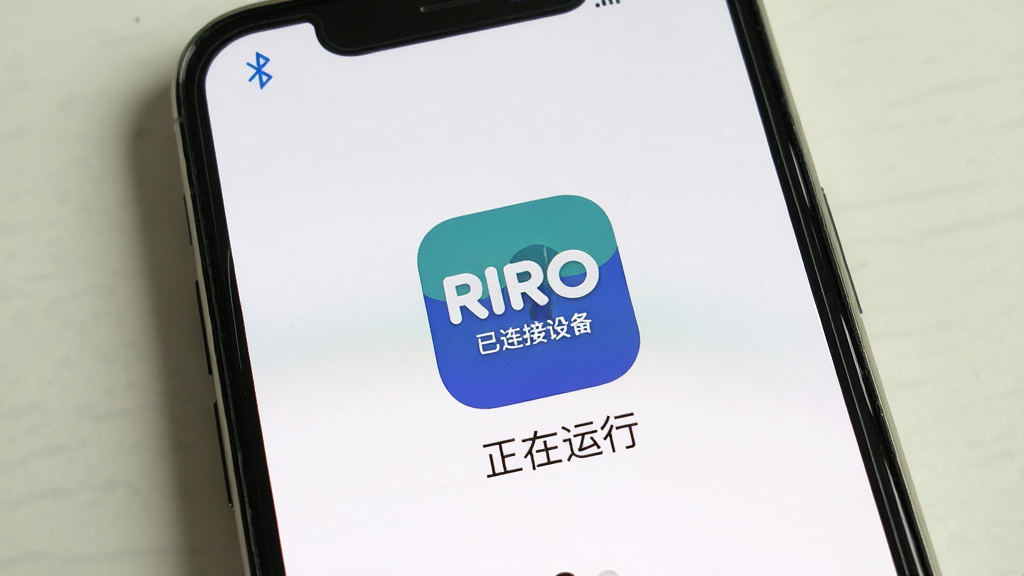主体:RIRO应用运行在手机上的实际效果,背景为纯色桌面。布局:手机屏幕占据画面主体,周围留白。排版:应用图标突出显示,屏幕下方有“正在运行”字样。风格:真实场景拍摄风格,光线柔和。细节:手机顶部状态栏显示蓝牙图标,屏幕内显示“RIRO 已连接设备”。画质:清晰自然,无多余元素。