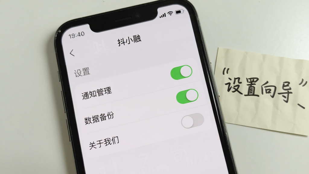 手机屏幕特写,显示“抖小融”设置页面,包含“通知管理”、“数据备份”、“关于我们”等选项,页面背景为浅白色,顶部有灰色状态栏;旁边摆放一张纸条,写有“设置向导”字样;构图居中,风格简洁,色彩素雅;细节包括开关按钮的绿色高亮状态;画质干净,无多余元素。