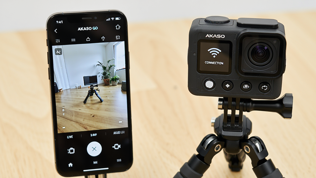 A smartphone displaying the AKASO GO app interface with a live camera preview feed, next to an AKASO action camera on a tripod, showing Wi-Fi connection status and control buttons, clean modern design, bright and clear UI elements, on a wooden desk background, professional product photography style, high resolution, sharp details, no clutter