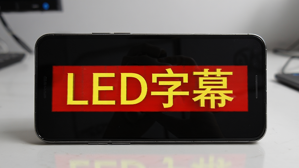 一部安卓手机横屏放置在桌面上,屏幕显示红底黄字的滚动字幕“LED字幕”,背景是简洁的室内桌面环境,主体突出手机屏幕内容,布局居中,风格写实科技感,细节清晰展示字幕发光效果与手机边框质感,画质高清,干净,无多余元素