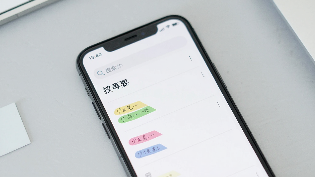 一张手机屏幕正面展示记事本应用界面的图片,画面中央是手机,手机屏幕上显示简洁的记事列表界面,顶部有搜索框,下方排列着几条彩色标签的笔记条目,背景为浅灰色桌面,整体风格清新简约,画质清晰干净,无多余元素
