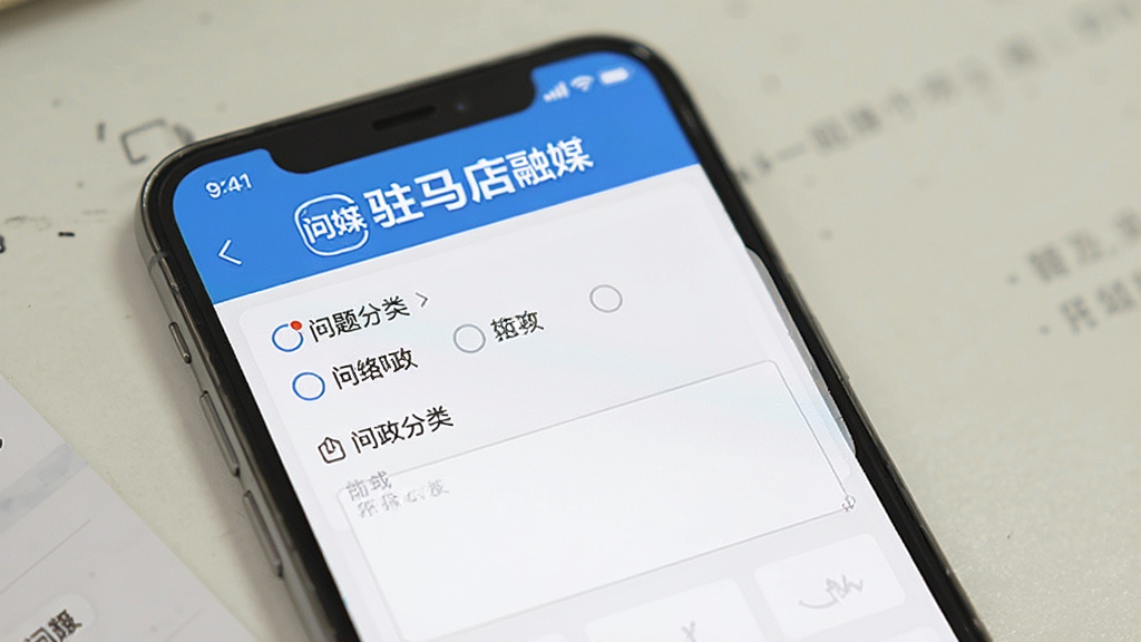 一张展示驻马店融媒网络问政功能界面的预览图,手机屏幕显示问政提交表单页面,包含问题分类选项和文字输入框,页面顶部有“驻马店融媒”文字标识,整体配色为蓝色和白色,风格正式简洁,画质干净,无多余元素