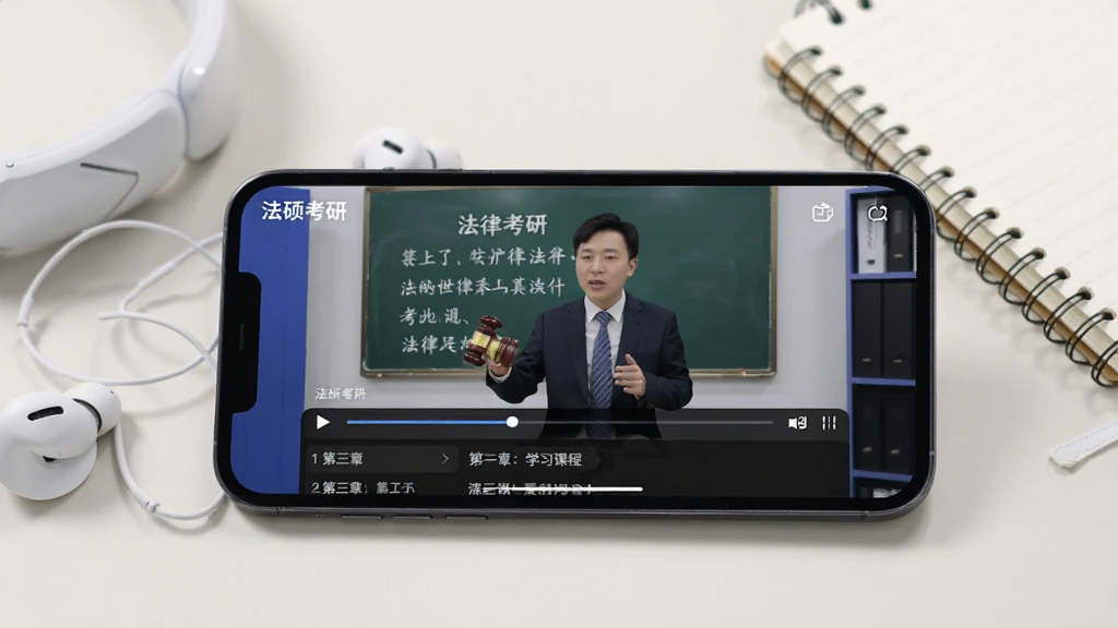 一张在线课程学习场景图,画面中一部手机横屏放置,屏幕显示“法硕考研”APP的课程播放界面,视频画面中有一位讲师正在讲解法律知识点,屏幕下方有进度条和章节列表。手机旁边有一副白色耳机和一本笔记本。背景为浅色桌面,风格简约现代,画质清晰,无多余元素。
