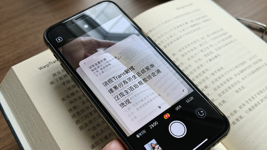 一张展示 WarpTrans 拍照翻译功能的图片,画面中手机摄像头对准一本外文书籍,屏幕上叠加着翻译后的中文浮层,主体为手机与书籍,布局对角线构图,排版自然,风格生活化写实,细节包含书页纹理与屏幕反光,画质清晰,干净,无多余元素