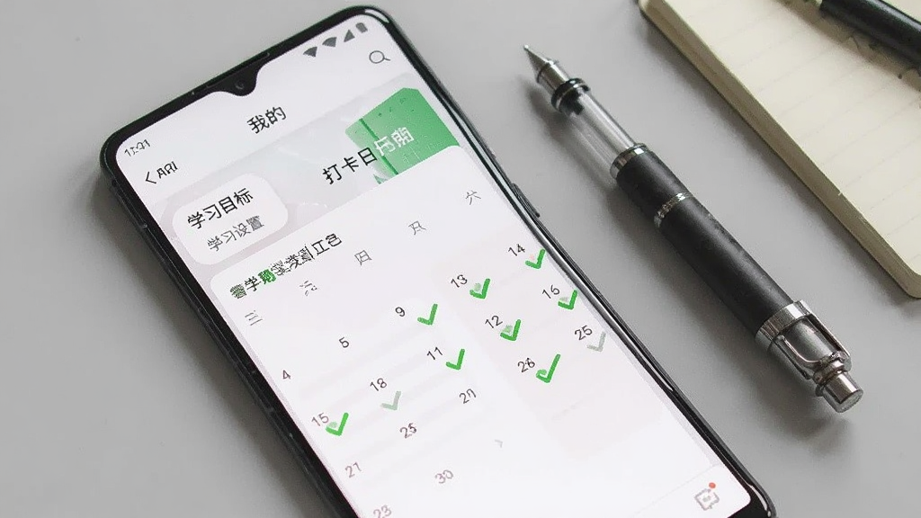 一部安卓手机显示日日新APP的“我的”页面,页面上有学习目标设置入口和打卡日历,日历上多个日期显示已打卡的绿色圆点,手机旁边放着一支笔和小本子,背景是浅灰色桌面,整体色调柔和,画面干净整洁