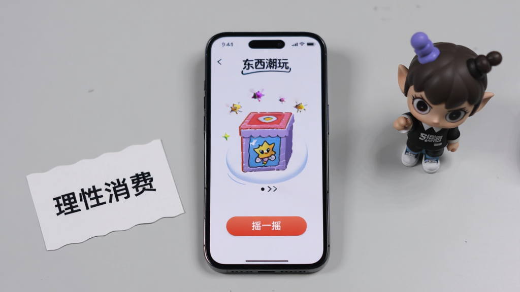 画面中央是一部智能手机,手机屏幕上显示“东西潮玩”的抽盒页面,页面中有“摇一摇”按钮和盲盒动画效果。手机旁边放着一张写有“理性消费”的提示卡片和一个迷你潮玩公仔。背景是浅灰色的桌面,整体布局采用手机居中、物品环绕的构图,风格为写实产品摄影,细节上突出手机屏幕的清晰度和公仔的质感。画质高清,画面干净,无多余元素。