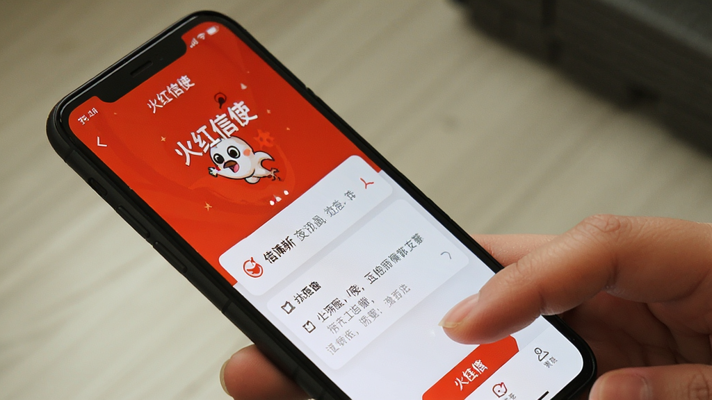一张火红信使在手机上运行的实拍风格示意图,主体为手持手机展示“火红信使”应用界面,布局采用斜角拍摄视角,风格为真实生活场景,细节包含手指点击屏幕和清晰的UI元素,画质高清干净无多余元素