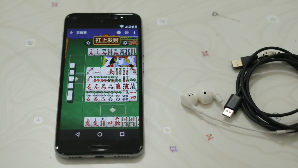 一部安卓手机显示“杠上发财”游戏对局进行中的画面,牌面清晰,界面整洁,手机旁边放着耳机和充电线,背景是简洁的桌面,风格生活化,布局自然,画质清晰,干净无多余元素