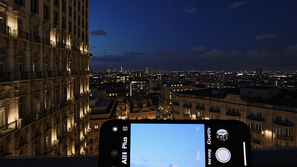 画面展示一张夜景城市照片,灯光璀璨,建筑细节清晰,天空呈现深蓝色。画面下方放置一部手机,屏幕显示AI相机Plus的拍摄界面,整体布局为照片占据大部分画面,手机位于右下角,风格写实,画质锐利,干净无多余元素。