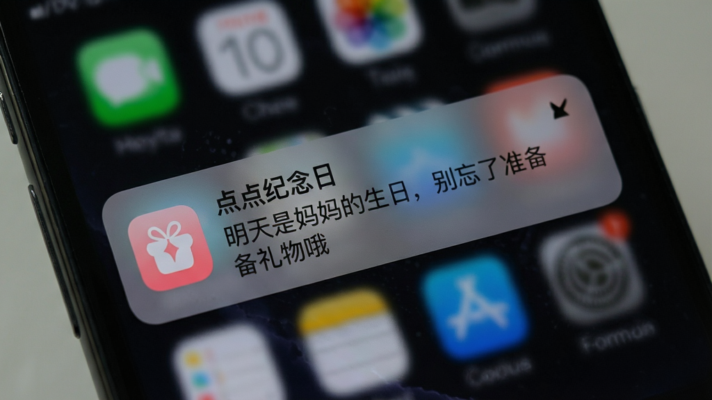 一部手机锁屏界面上显示“点点纪念日”推送通知的图片,通知内容为“明天是妈妈的生日,别忘了准备礼物哦”,通知卡片带有应用图标,背景为手机深色锁屏壁纸,画面聚焦于通知卡片,风格写实清晰,画质干净,无杂乱信息