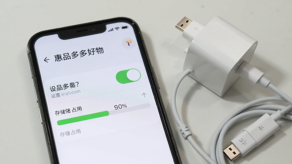手机屏幕显示“惠品多多好物”设置与权限管理界面,画面中有开关按钮和存储占用条,背景为浅色简洁风格,手机旁放着一个充电头和数据线,构图清晰,画质干净,无多余元素