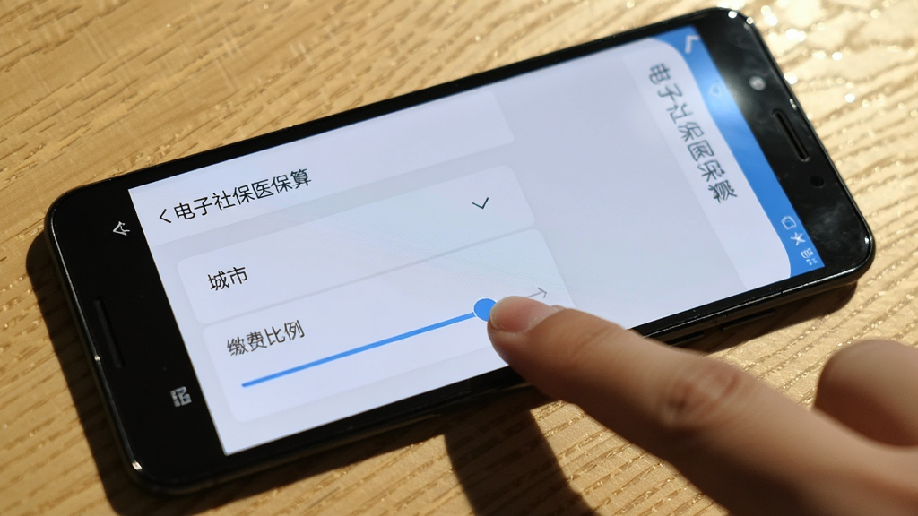 一部安卓手机放在桌面上,屏幕显示“电子社保医保算”的设置页面,页面中有城市选择下拉菜单和缴费比例滑块,旁边有一只手正在点击屏幕,背景是木质桌面,风格为生活写实风,细节包括手指阴影和屏幕反光,画质清晰,干净,无多余元素