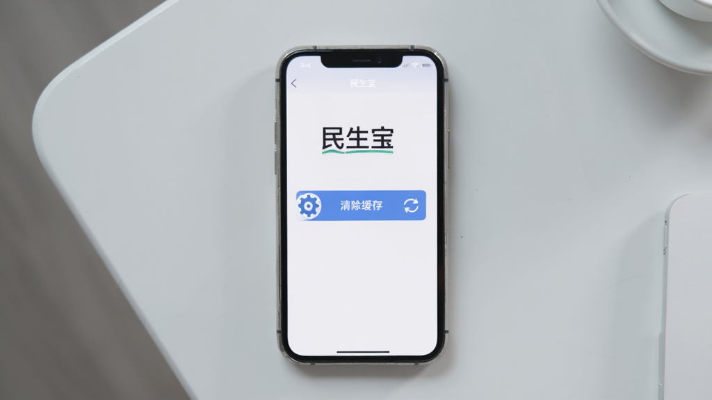 画面展示一部手机放置在桌面上,屏幕显示民生宝设置界面中的“清除缓存”按钮,旁边有齿轮图标和刷新箭头,背景为浅灰色简约桌面,布局居中,风格干净清爽,细节上突出“民生宝”文字与设置图标,画质清晰,无杂乱元素