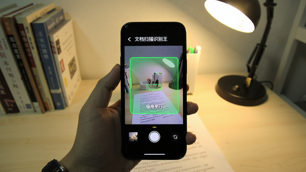 画面主体为一只手拿着手机正在拍摄一份纸质文档,手机屏幕上显示