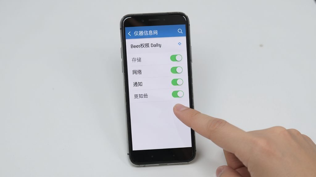 仪器信息网新手安装设置界面展示图,画面主体为一部竖直放置的智能手机,屏幕内显示应用权限设置页面,包含存储、网络、通知等权限开关,手机旁边有一只手正点击屏幕,背景为浅灰色,风格教程指引类,画质清晰,干净无多余元素