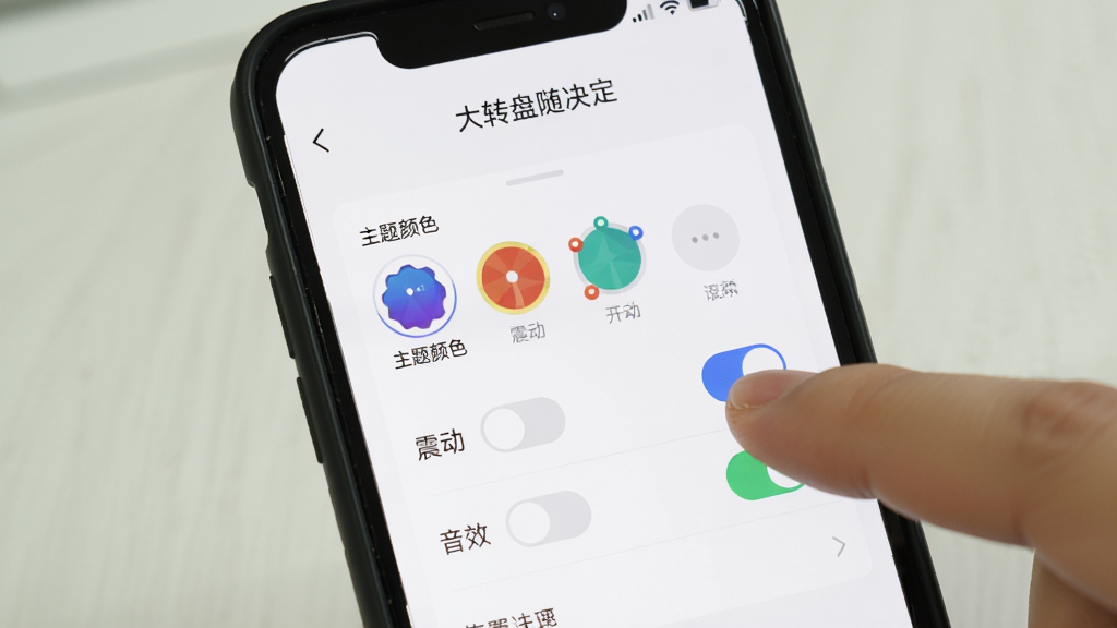 手机屏幕显示“大转盘随决定”的设置页面,页面上有主题颜色选项、震动开关和音效开关,用户手指正在点击其中一个开关按钮,界面背景为白色,风格清爽,画质清晰,干净无多余元素
